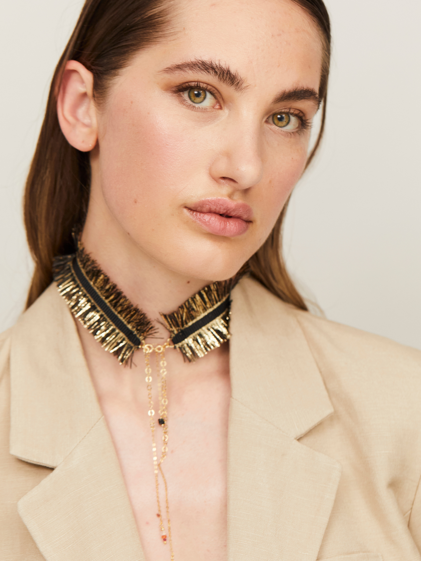 Exzentrische Halskette in Schwarz und Gold, getragen von einer jungen, attraktiven Frau, die zusätzlich einen hellen Blazer trägt und in die Kamera guckt.