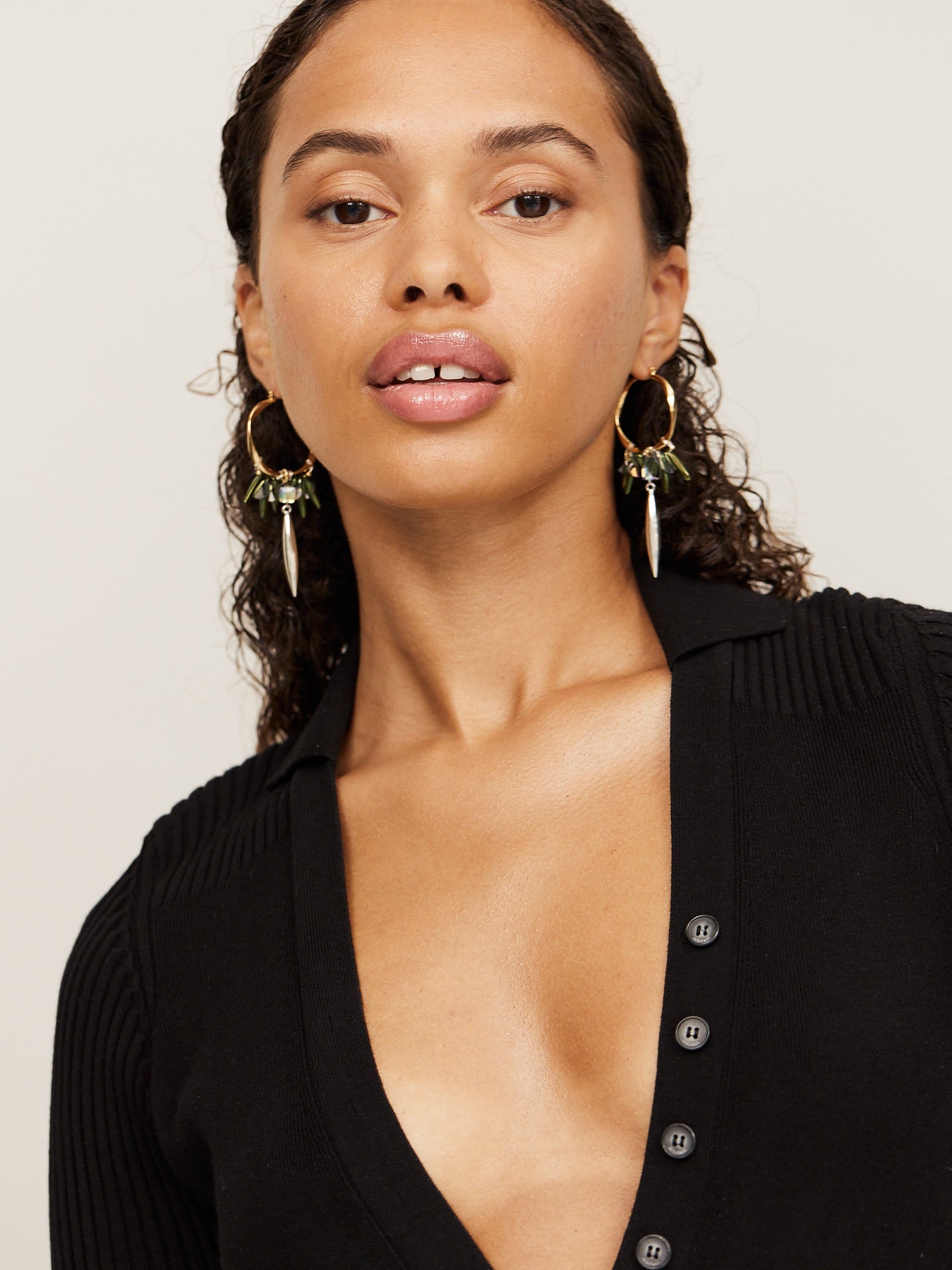 Attraktive Frau mit schwarzen Haaren und dunklen Augen trägt einen schwarzen Body und goldene Kreolen mit grünen Steinen und einem silbernen langen Anhänger.