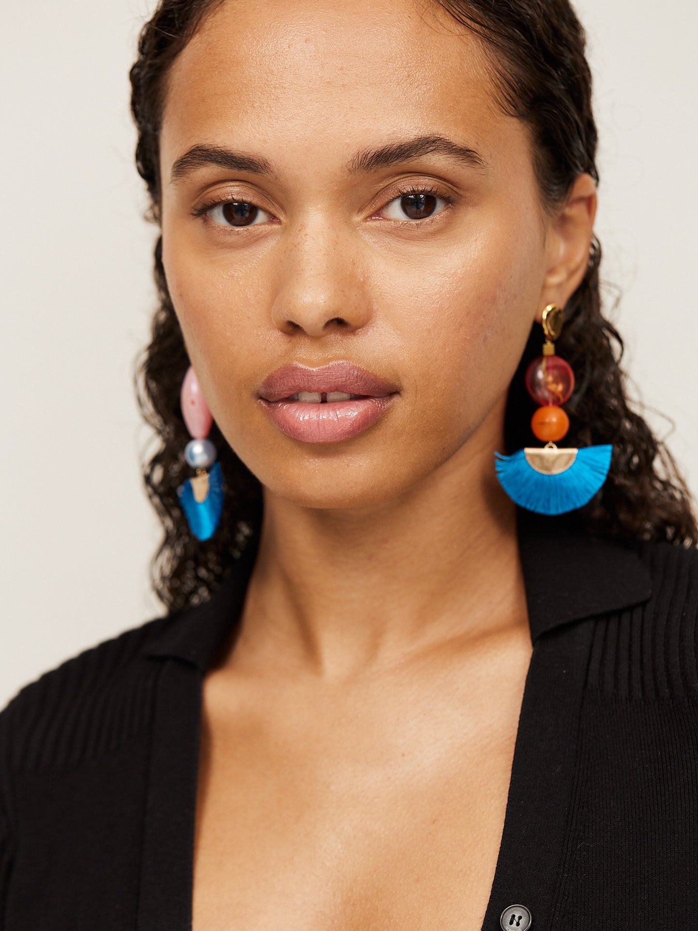 Attraktive Frau mit braunen Augen trägt einen schwarzen Body und aufregende Ohrringe mit einem tollen blauen Fächer und einer orangefarbenen Perle. Der asymmetrische andere Ohrringe ist farblich ähnlich, in seiner Form aber etwas länger.