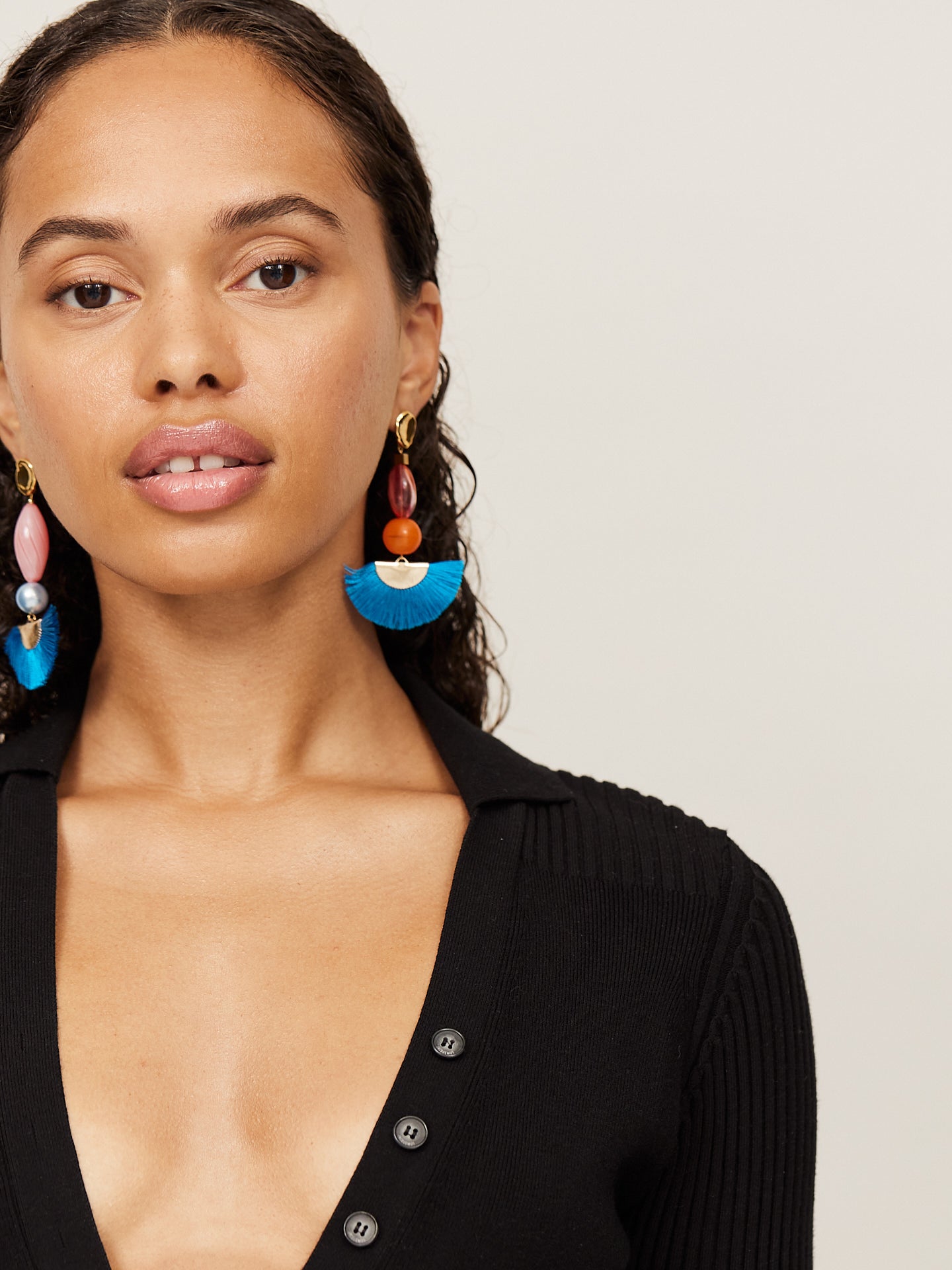 Attraktive Frau mit braunen Augen trägt einen schwarzen Body und aufregende Ohrringe mit einem tollen blauen Fächer und einer orangefarbenen Perle. Der asymmetrische andere Ohrringe ist farblich ähnlich, in seiner Form aber etwas länger.