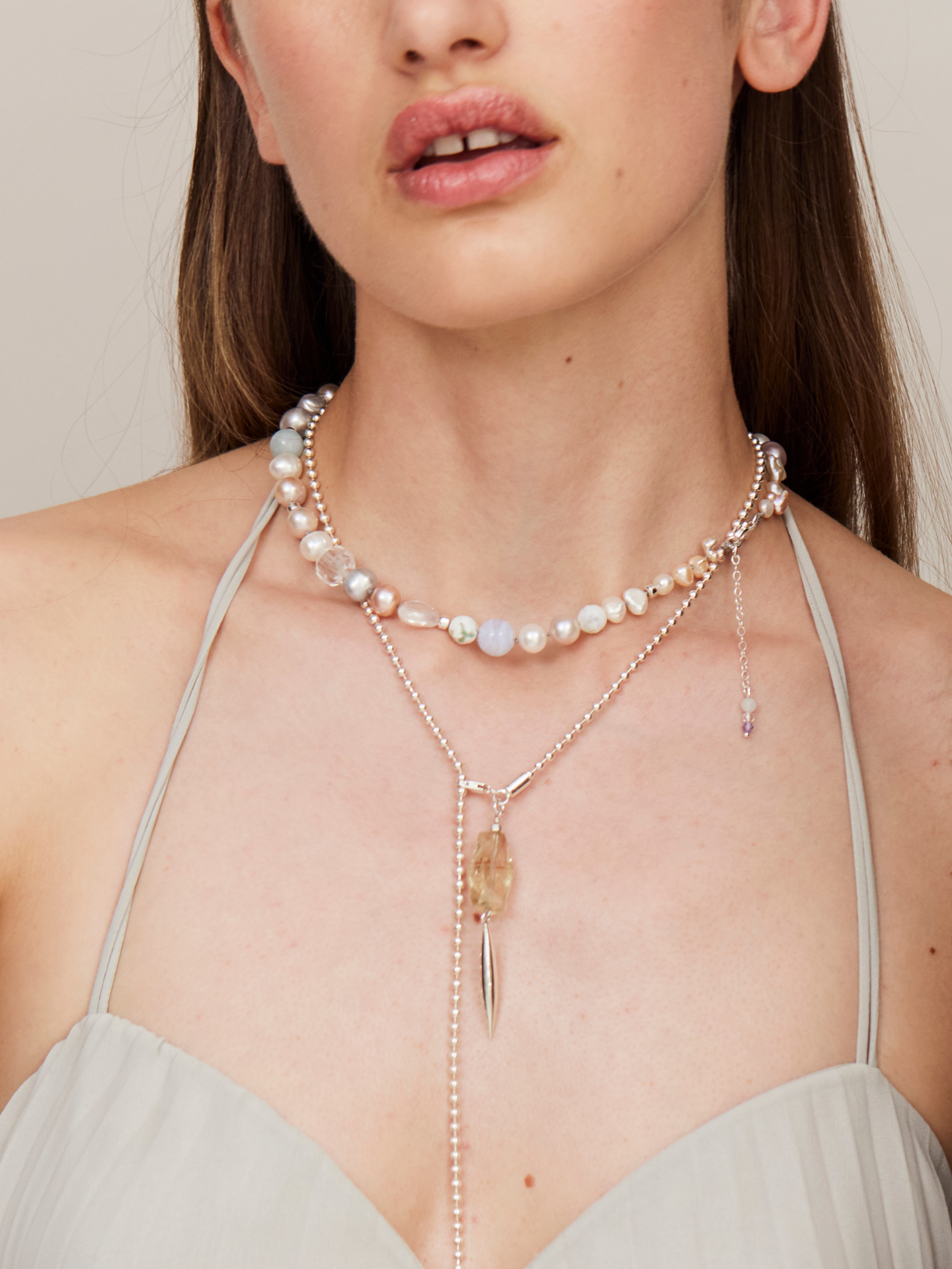 Attraktive Frau mit sinnlichen Lippen trägt ein helles, schulterfreies Kleid und eine Kombi aus einer Perlenkette und einer zarten Halskette mit einem aufregenden Anhänger